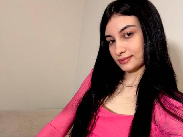 DevineSimone has long black hair, pink top, and a subtle smile. She's sitting with a slight smirk, showing her tits and a small nose piercing.