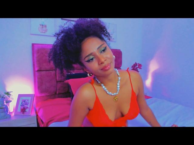 MeganWilliams with curly black hair, wearing a red dress with a low neckline, showing her tits, a white bead necklace, and a gold pendant, sitting on a bed.