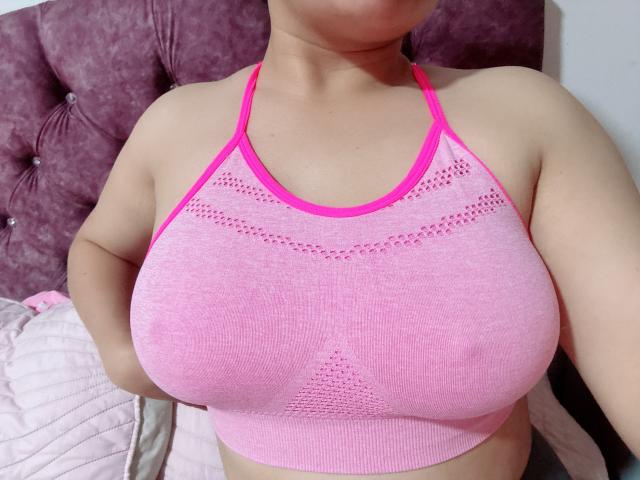 MiaDuboisX, com seios grandes, vestindo um top esportivo rosa de malha, com alças finas, mostrando seus mamilos rosa através do tecido. Ela está sentada em uma cama com uma cabeceira rosa..