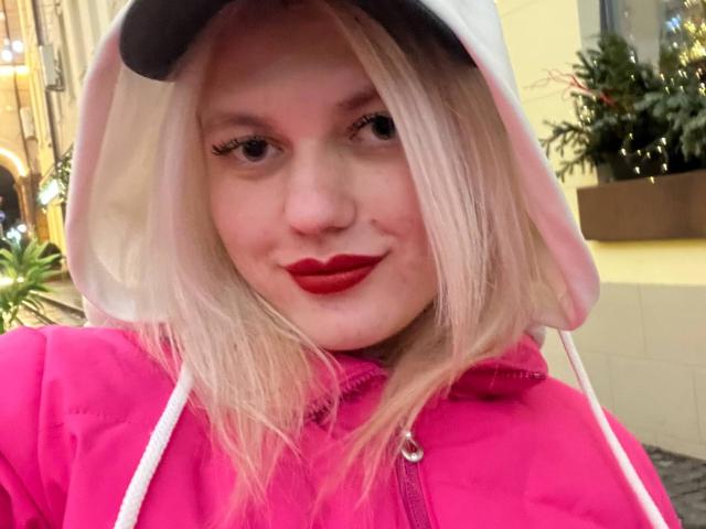 ToniField tiene el cabello rubio, lápiz labial rojo, una sudadera rosa y una capucha blanca con orejas negras, y está sonriendo con una expresión linda y ligeramente traviesa. Sus senos no son visibles..