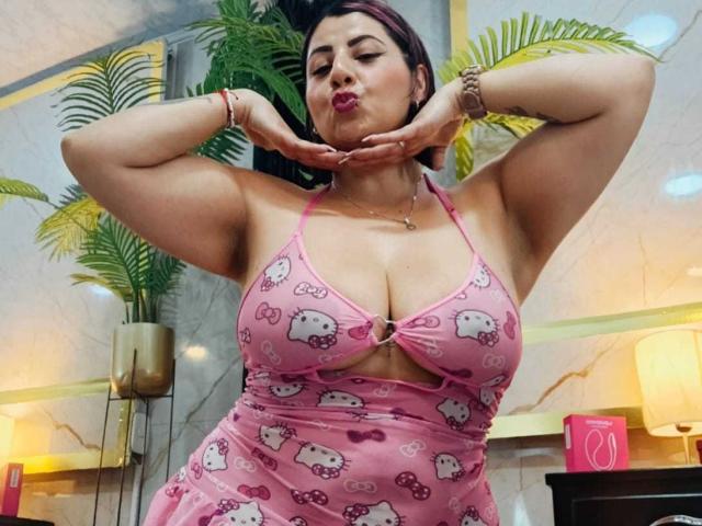 AngeleRouxo com seios grandes, vestindo um vestido rosa do Hello Kitty, fazendo beicinho com as mãos perto do queixo, em pé em um quarto com plantas verdes e um abajur rosa..