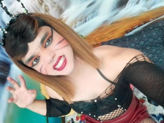 GabiiSexy, avec ses cheveux orange-brun, son haut en résille noir, sa jupe rouge et son maquillage avec un motif de chat, sourit et fait signe de la main, montrant de petites seins et un visage mignon et sexy..