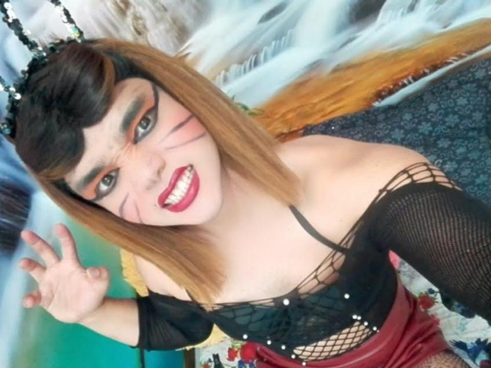 GabiiSexy with orange-brown hair, black fishnet top, red skirt, and catface makeup, smiling and waving, showing small tits and a cute, sexy face.