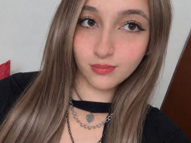 MiluSantoro hat langes, braunes Haar, braune Augen mit schwarzem Eyeliner, rosafarbenen Lippenstift, eine schwarze Halskette mit silberner Kette und ein schwarzes Oberteil. Sie schaut mit einem leichten Lächeln in die Kamera..