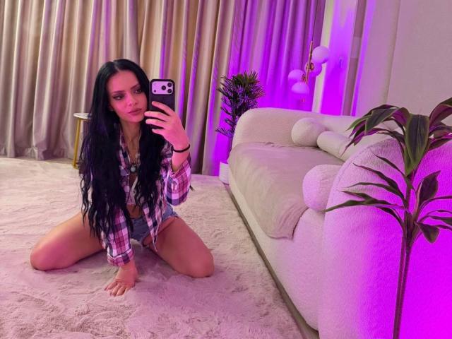 IliriaKara, avec ses longs cheveux noirs, sa chemise à carreaux et son short en jean, est agenouillée sur un tapis rose, en train de prendre un selfie devant un miroir avec son téléphone, montrant ses parties intimes. L'éclairage est violet..