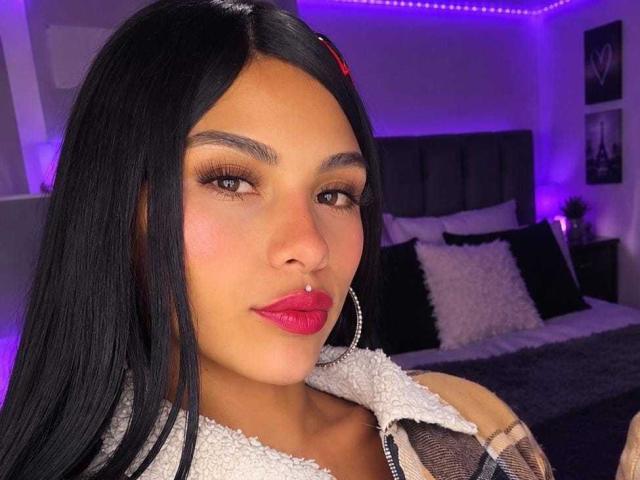 ClarisCampbell has long black hair, brown eyes, red lipstick, a nose ring, and hoop earrings. She's wearing a white, furry, sleeveless top. Her ass is not visible.