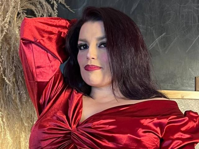 SachyMoon, com cabelos longos e escuros, vestido de cetim vermelho, mangas bufantes e batom vermelho, braços levantados, exibindo os seios, com uma expressão confiante e sexy..
