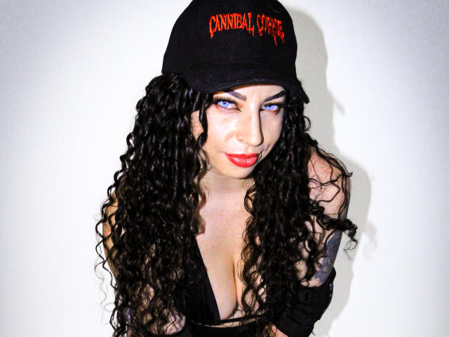 ScarlattaaDesire with long curly black hair, blue eyes, red lipstick, black cap, and deep cleavage in a black top, looking up with a slight smile.