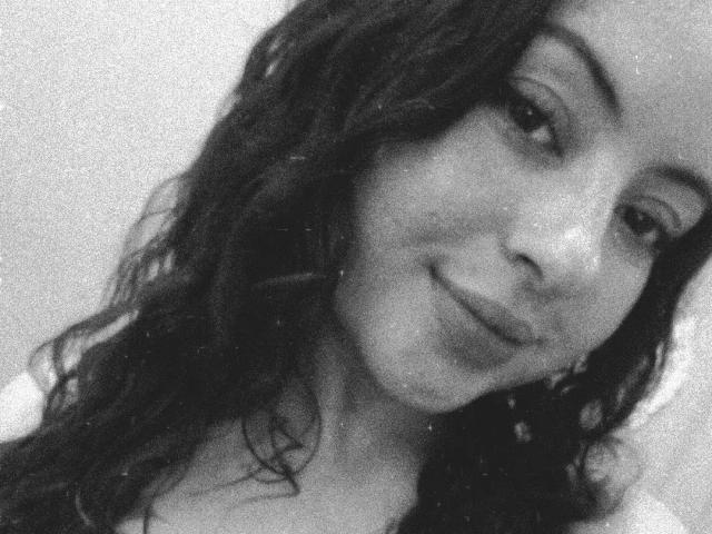 Autoportrait en noir et blanc, légèrement flou, de MiiaBennet avec des cheveux noirs ondulés, un léger sourire et un petit piercing au nez. Ses seins sont partiellement visibles. Elle a l'air mignonne et détendue..