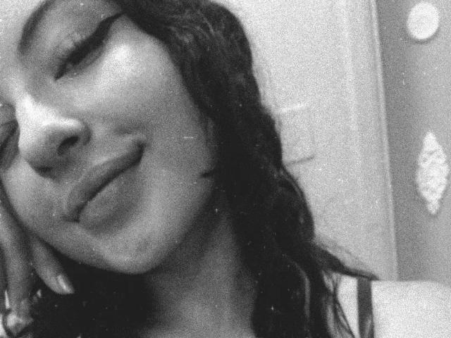 Grainy black-and-white selfie of MiiaBennet with her head tilted to the side, smiling slightly, showing a small nose and full lips. She has long, wavy hair and is touching her cheek.