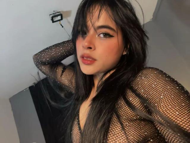 SophieBanksX, met lang zwart haar, draagt een glinsterende fishnet top, waarbij haar borsten zichtbaar zijn. Ze heeft haar arm achter haar hoofd en kijkt naar de camera met roze lippenstift..