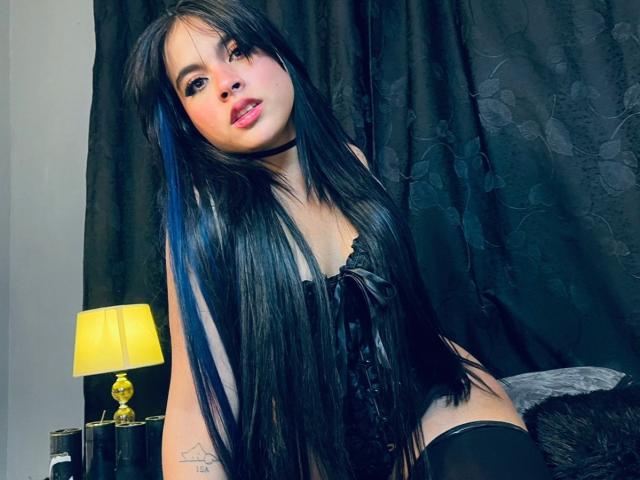 SophieBanksX, avec de longs cheveux noirs et des reflets bleus, portant de la lingerie noire et des bas hauts, assise, avec un fond de rideau noir, une lampe jaune, regardant la caméra avec des lèvres roses, tatouage sur le bras..