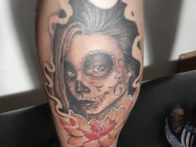 SandraXSeins' tattoo on a leg shows a detailed, black-and-gray sugar skull with dark eyes, a stitched mouth, and flowing hair, surrounded by flame-like patterns and red flowers.