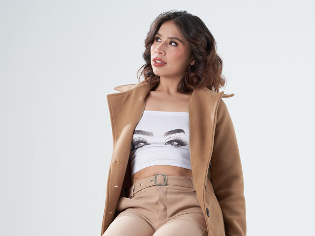 MacarenaJonesFg with brown wavy hair, tan coat, white eye-print crop top, and beige pants, looking to the side, showing her tits slightly through the top.
