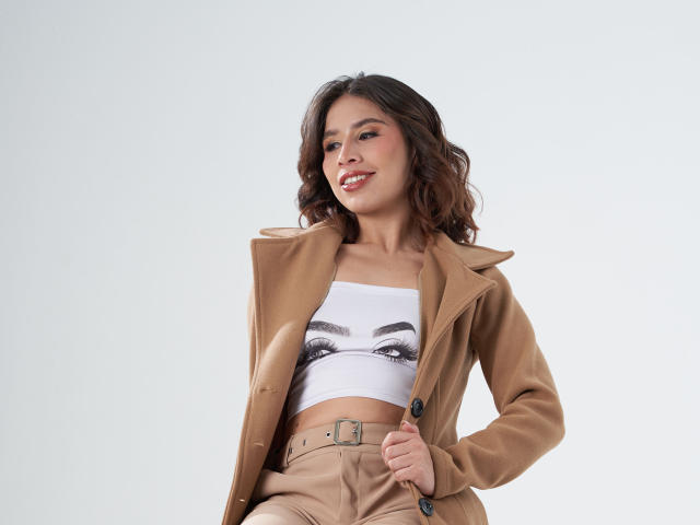 MacarenaJonesFg met golvend bruin haar, lachend, gekleed in een beige jas over een witte top met een opdruk van ogen, en beige broek, terwijl ze een knoop van de jas vasthoudt, waarbij haar borsten zichtbaar zijn..