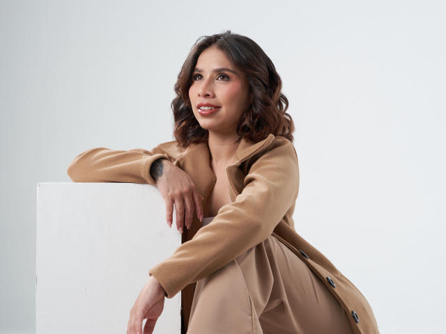 MacarenaJonesFg, donna con capelli castani e ondulati, cappotto color sabbia e gonna, appoggiata a un cubo bianco, sorridente, che mostra piccoli seni, carina, sexy, pelle chiara, scatto in studio..