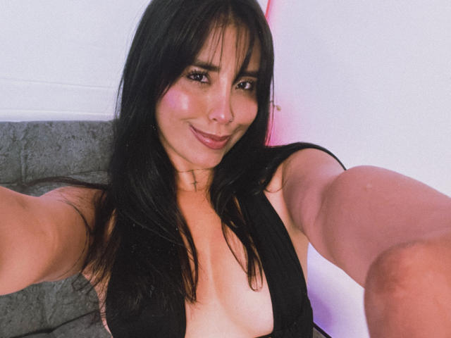 Elizabeth Macy, avec de longs cheveux noirs, souriante, portant un haut noir à décolleté plongeant qui révèle ses seins, prenant un selfie en étendant son bras, avec un éclairage rose et blanc en arrière-plan..