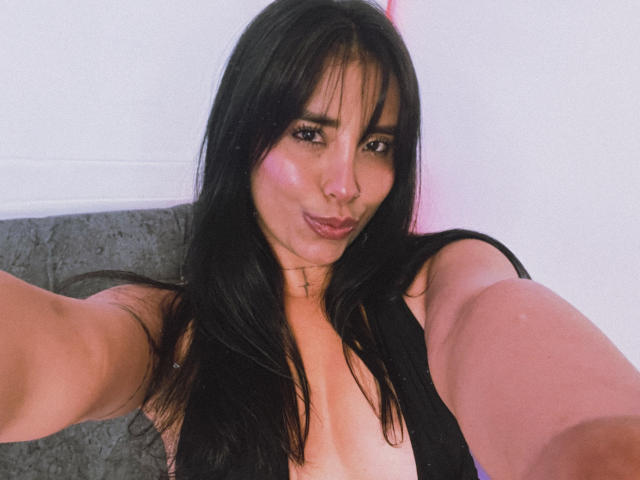 Elizabeth Macy, com cabelos pretos longos, olhos castanhos e um sorriso adorável, vestindo uma blusa preta, mostrando seus seios, tirando uma selfie, com os braços estendidos, usando um colar com uma cruz..