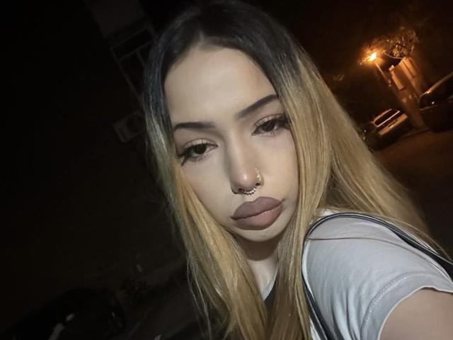 IvyGlowX tiene el pelo largo y rubio, un piercing en el tabique nasal y labios carnosos y sensuales. Lleva una camisa blanca con tirantes negros y mira hacia abajo con una expresión seria..