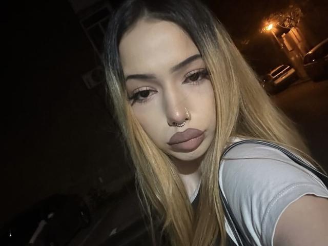 IvyGlowX with long blonde hair, nose ring, and full lips, wearing a gray shirt, looking at the camera with half-closed eyes in a dimly lit, dark background.