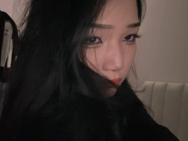 AricaWings has black hair, dark eyes, and slightly parted red lips with a small piercing on the bottom right. Her expression is subtle, looking to the side.