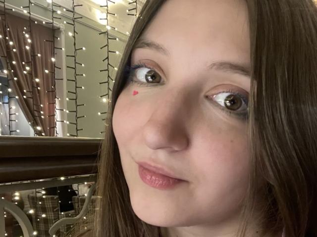 PhuongFagnan's face, brown eyes, light skin, small pink heart on cheek, blue eyeshadow, brown hair, slightly smiling, glossy pink lips, soft lighting, warm indoor setting.