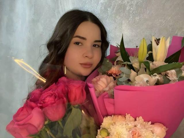 NoelZeiler, con i capelli scuri, tiene in mano un bouquet rosa di rose, gigli e crisantemi, sorridendo alla fotocamera, con un bengala in primo piano..