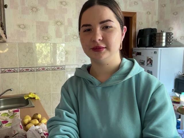 NoelZeiler, met een lichte huid, bruin haar in een staart, draagt een turquoise hoodie, heeft roze lippenstift, kleine oorbellen en een lichtjes fronsende uitdrukking, en staat in een keuken..