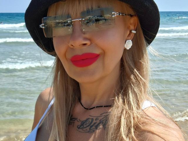 HotAdelayde69 with bright red lips, wearing a black hat, reflective sunglasses, white top, and a necklace, has a tattoo on her chest, and is at the beach.