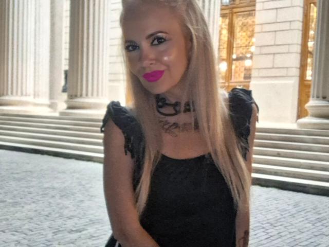 HotAdelayde69 with long blonde hair, black lace dress, pink lipstick, black choker, standing on cobblestone, smiling, showing off her tits, sexy, cute, attractive, in front of building.