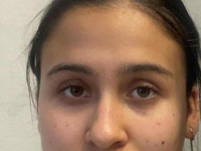 JamesPaino's face, showing brown eyes, slightly arched eyebrows, small nose, and a few dark moles on their fair skin. They have dark hair pulled back, wearing a gold earring.