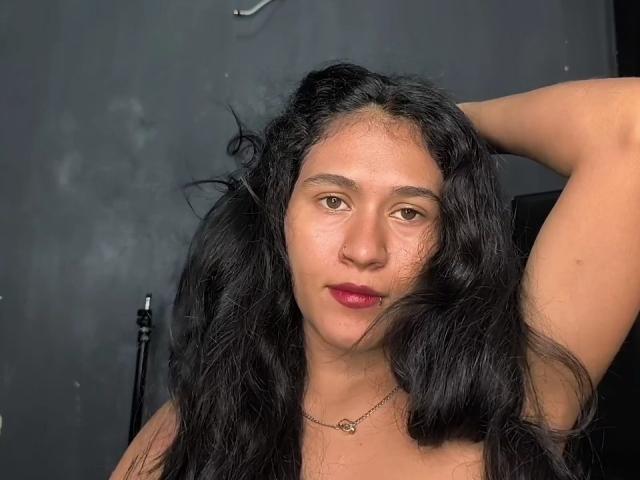 Angelicap69 has long black wavy hair, brown eyes, and a nose ring. She's raising her right arm, showing her bare shoulder, with a necklace and red lipstick.