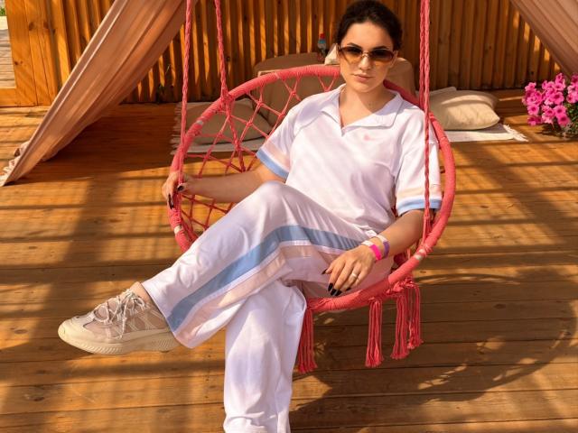 Dreia with dark hair, sunglasses, and white shorts with blue stripes, sitting in pink macramé swing, wearing beige sneakers, wooden deck, pink flowers, and wristbands.