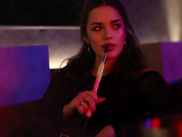 Dreia with dark hair, black dress, holding a glowing pink and purple bong to her mouth, looking to the side with a slight smirk. Her nails are painted red.