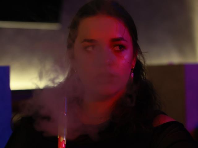 Dreia with smoky face, red and purple light reflecting on her, dark wavy hair, black top, small earrings, slightly blurred, looking to the side.