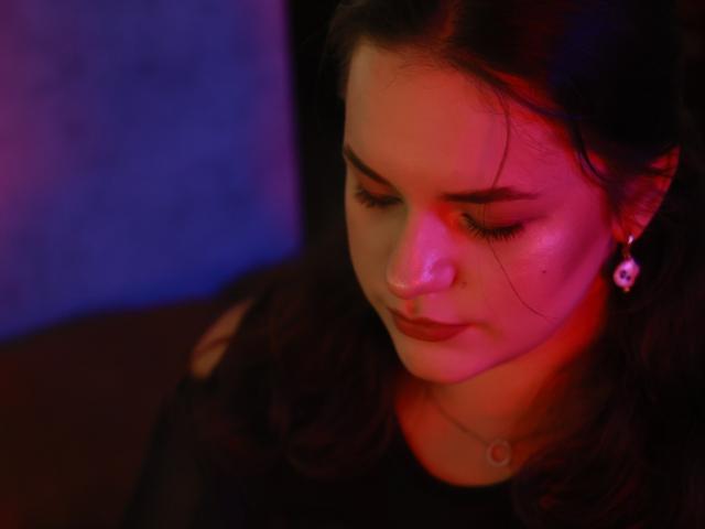 Dreia a les cheveux foncés, des lumières rouges et violettes sur son visage, les yeux fermés, et une expression pensive. Elle porte un haut noir avec un petit pendentif, et un boucle d'oreille en diamant..