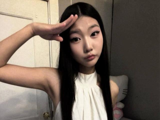 MaragretWilmeth a de longs cheveux noirs, elle porte un haut blanc sans manches, elle salue de la main droite et a une expression mignonne et sérieuse. Ses petites seins sont visibles..