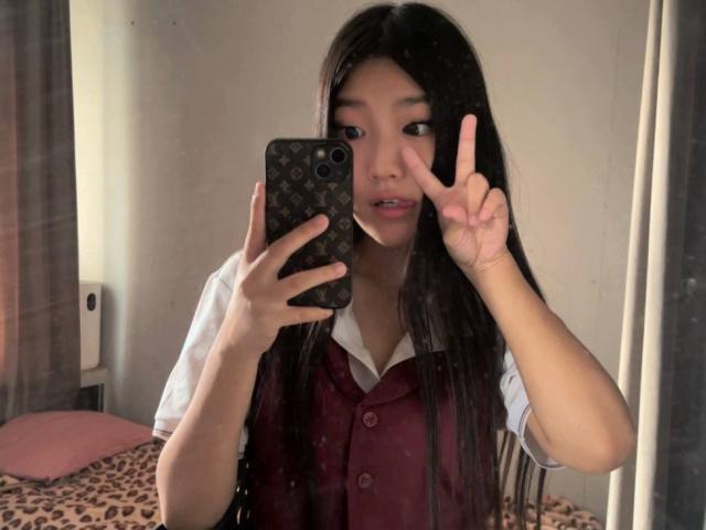 Maragret Wilmeth, avec de longs cheveux noirs, portant un gilet bordeaux par-dessus une chemise blanche, faisant un signe de paix de la main droite, prenant un selfie devant un miroir avec son téléphone..