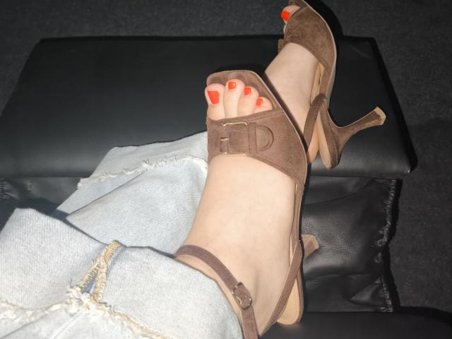 Les pieds de Dreia, avec du vernis à ongles rouge, portant des escarpins marron en daim, reposent sur un tabouret en cuir noir, les jambes en jean bleu clair, avec une bride visible à la cheville de la chaussure..