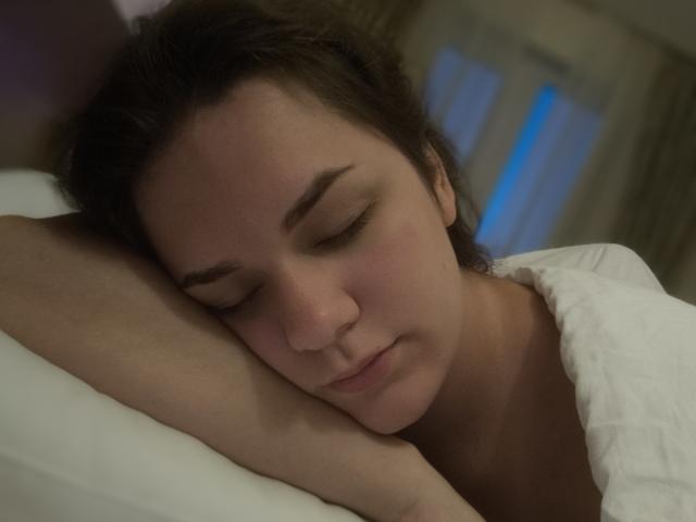 Le visage de Dreia est paisible, les yeux fermés, les sourcils bruns légèrement relevés, et la tête reposant sur un oreiller blanc. Elle a les cheveux bruns et une couverture blanche..