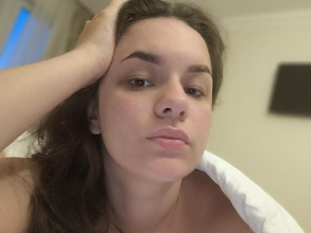 Dreia, avec sa main dans ses cheveux bruns, a l'air légèrement ennuyée, a des lèvres rosées et des yeux marron, porte une chemise blanche, et a un teint clair et un petit nez..
