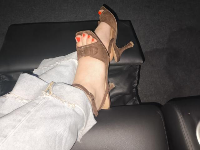 Dreia's foot in brown, suede, high-heel sandal with her red-painted toes, resting on black leather, showing the ankle of her ripped, light-blue jeans.