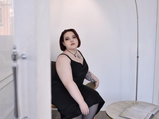 IvoryeFlame, a curvy woman with pale skin and red bob, sits in a black dress, showing a tattoo on her arm, with a chair and lamp in the room.