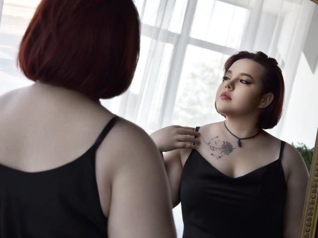 IvoryeFlame with red bob, black tank top, tattoo on chest, looking at reflection, touching shoulder, black necklace, pale skin, soft light, mirror, slightly pouty expression.