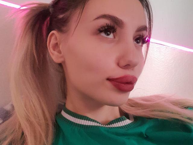 OtakuOops con coletas rubias, camisa verde, lápiz labial rojo y pestañas largas, mirando de lado con una ligera sonrisa, luz neón rosa de fondo..
