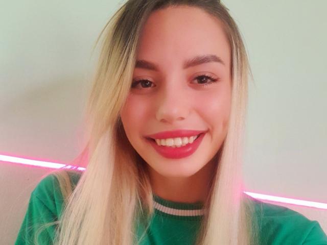 A OtakuOops tem cabelo loiro, olhos verdes e batom vermelho, está sorrindo mostrando os dentes brancos, vestindo uma camisa verde, e uma luz neon rosa está ao fundo..