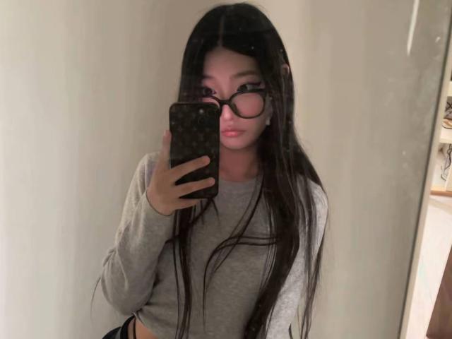 Maragret Wilmeth mit langem, schwarzem Haar, die eine Brille trägt und ein graues, langärmeliges Hemd anhat, macht ein Selfie mit ihrem Handy und zeigt dabei ihre flachen Brüste und einen leichten Schmollmund..