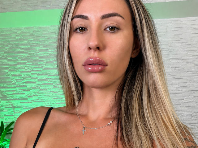 VanessaSky heeft glanzende, volle lippen en lang, steil blond haar met donkere aanzet. Ze draagt een zwarte bandeau en een delicate kruisbedel. Haar ogen zijn licht geopend..