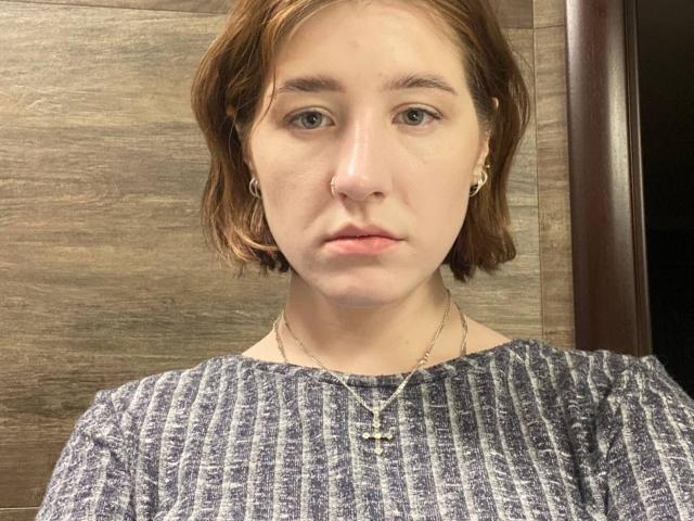 Malvina Hauenstein, con cabello castaño corto, vestida con una camiseta gris de canalé, un collar delgado y pequeños aretes, tiene una expresión neutra y piel pálida. No se ven los senos..