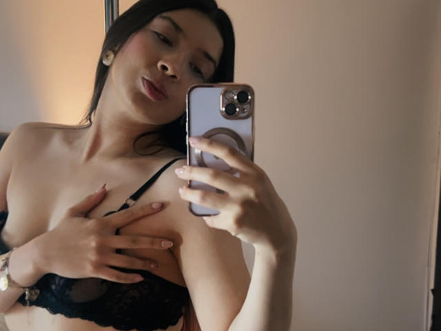 EmmaLombardi taking a mirror selfie, wearing a black lace bra, covering her tits with one hand, holding a phone with a ring design in the other.